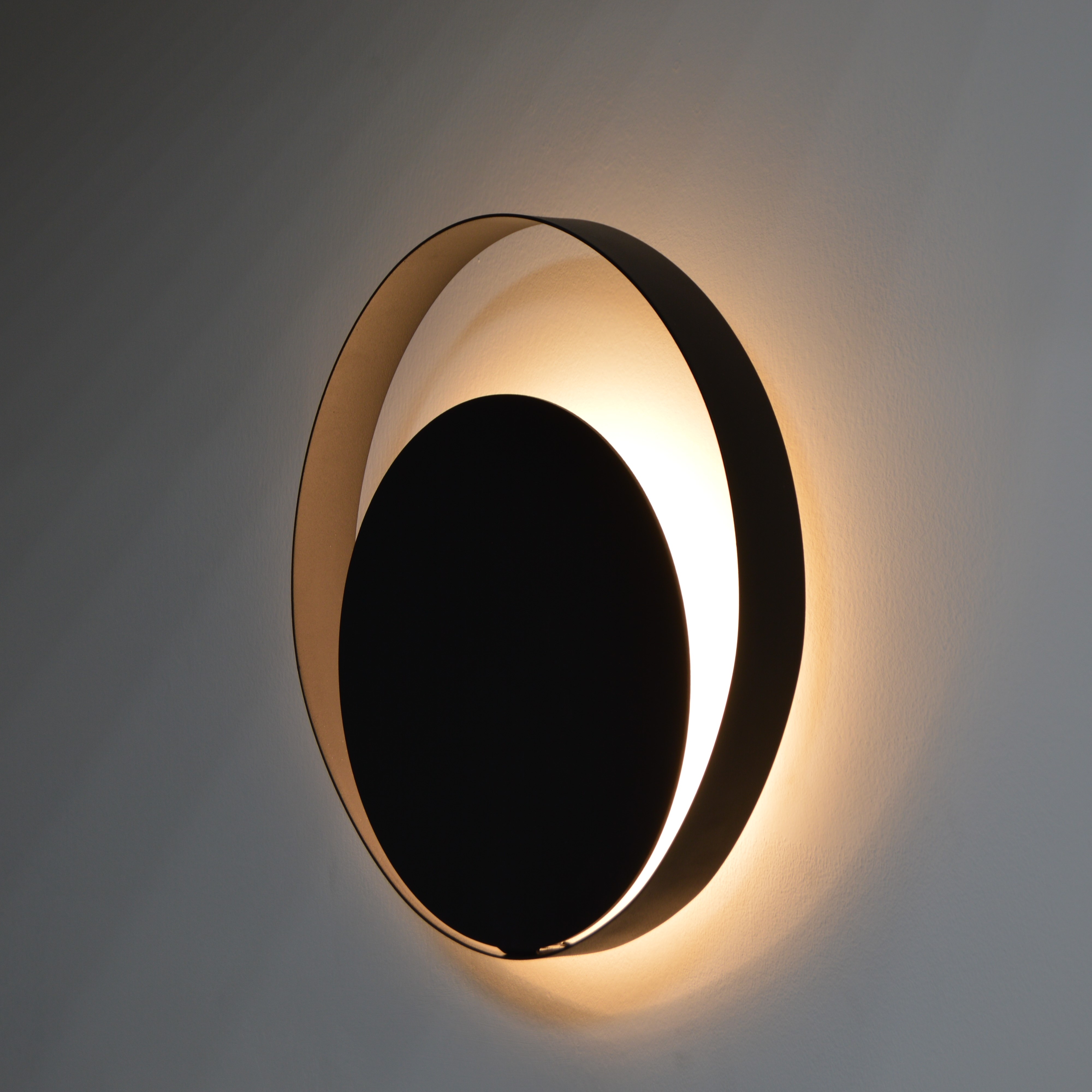 Luminária de parede resistente à água modelo Eclipse Ø40cm instalada em área externa com parede cinza, poltrona azul e plantas decorativas.